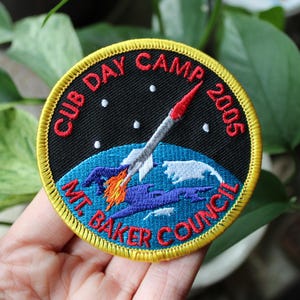 May include: Embroidered patch with a rocket ship design. The patch features the text "CUB DAY CAMP 2005" in red, and "MT. BAKER COUNCIL" in red. The rocket ship is white and red, with blue and orange flames. The patch has a yellow border.
