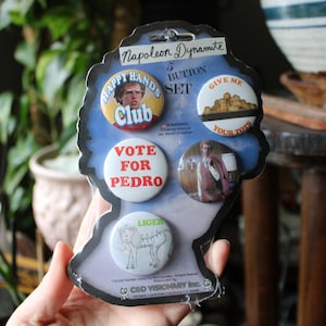 May include: A button set featuring images and text from the movie "Napoleon Dynamite." The buttons include phrases like "Happy Hands Club," "Vote for Pedro," and "Give Me Your Tots." The set is packaged on a blue and black backing.