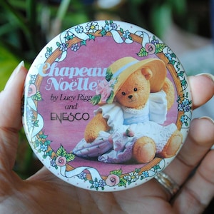 May include: A round, pink and white compact mirror with a picture of a teddy bear wearing a hat and a dress. The mirror has a white ribbon with the text "Chapeau Noelle by Lucy Riggs and ENESCO" printed on it.
