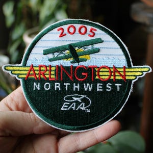 May include: Embroidered patch with a green and white circular design. The patch features the text "2005", "ARLINGTON NORTHWEST", and an image of a biplane. The EAA logo is also present. The patch has yellow and red accents.