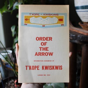 May include: A vintage handbook titled "Order of the Arrow" is held in front of a blurred background. The cover is cream-colored with red text and a blue and yellow emblem. The text includes "T'KOPE KWISKWIS" and "Lodge No. 502".