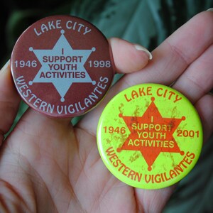 Op de afbeelding: Twee ronde speldjes. Eén is bordeauxrood met een witte ster en tekst: "Lake City, I Support Youth Activities, Western Vigilantes, 1946, 1998." De andere is geel met rode tekst: "Lake City, I Support Youth Activities, Western Vigilantes, 1946, 2001."