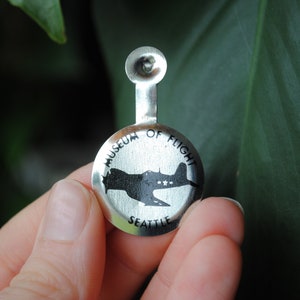 May include: Silver pinback button with a black and white silhouette of a plane and the text "Museum of Flight Seattle".