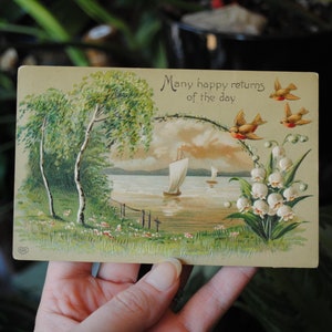 May include: Vintage postcard with a scenic illustration. The image features a lake scene with sailboats, trees, and a floral arrangement. The text "Many happy returns of the day" is written at the top, with birds flying above.