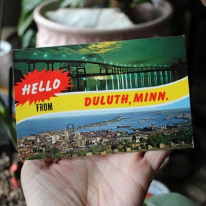 May include: Vintage postcard featuring a scenic view of Duluth, Minnesota. The postcard has a bridge over water at night and a cityscape during the day. Text on the postcard reads "HELLO FROM DULUTH, MINN."
