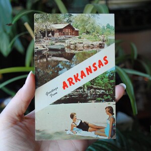 May include: A vintage postcard with a photo of a lake and a cabin in the woods. The postcard says "Greetings from Arkansas" in red letters.
