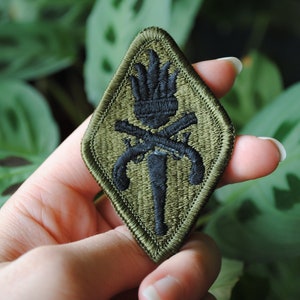 May include: A green diamond-shaped patch with black embroidered details. The patch features a torch with crossed rifles and a star at the top.