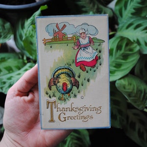 May include: Vintage Thanksgiving postcard featuring a girl in a red and white dress and a blue apron walking towards a turkey. The postcard has the text "Thanksgiving Greetings" in gold lettering.