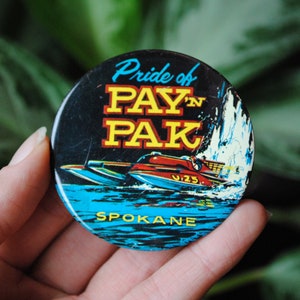 May include: A round button with a blue, white, and red image of a speedboat on water. The text "Pride of PAY'N PAK SPOKANE" is printed on the button.