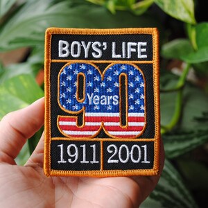 May include: A black and orange embroidered patch with the text "BOYS' LIFE 90 Years 1911 2001". The number 90 is designed with an American flag pattern.