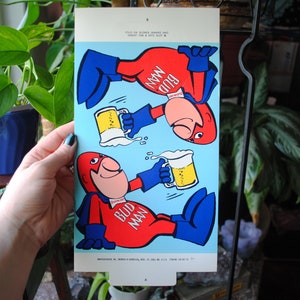 May include: A vintage Bud Man paper cutout with two images of a cartoon superhero holding a glass of beer. The superhero is wearing a red suit with a blue cape and blue boots. The text "Bud Man" is printed on the suit.