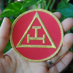May include: A round, red patch with a gold embroidered design. The design features a triangle with a horizontal line and a vertical line in the center, and a horizontal line below. The patch has a gold border.