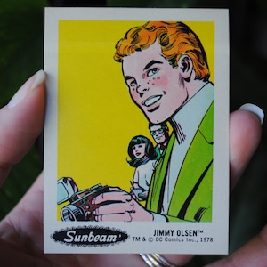 May include: A vintage comic book trading card featuring a cartoon illustration of a man with red hair and a green jacket holding a camera. The card is yellow with black text that reads "Sunbeam" and "Jimmy Olsen TM & © DC Comics Inc., 1978".