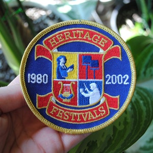 May include: A round, embroidered patch with a blue background and gold trim. The patch features the words "HERITAGE FESTIVALS" in red, with the years 1980 and 2002. The center has four squares with musical figures.
