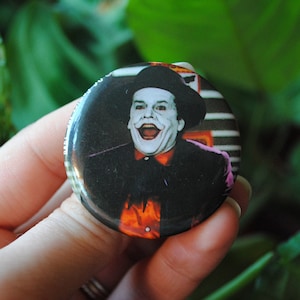 Puede incluir: Un botón redondo con una imagen en blanco y negro del Joker de la película de Batman. El Joker lleva un traje morado y un sombrero verde. Está sonriendo con una amplia sonrisa de dientes.