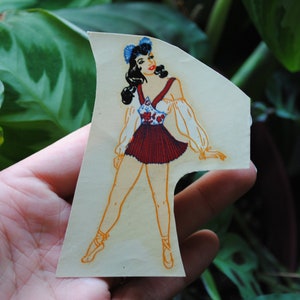 May include: A vintage paper doll cutout of a woman wearing a red and white dress with a blue bow in her hair. The doll is standing on a white background.