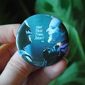 Puede incluir: Un botón redondo con una imagen azul y negra de Batman y el Joker. El texto en el botón dice "Not This Time, Joker!" con un símbolo de marca registrada.