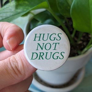 Puede incluir: Un botón blanco con texto verde que dice "HUGS NOT DRUGS".