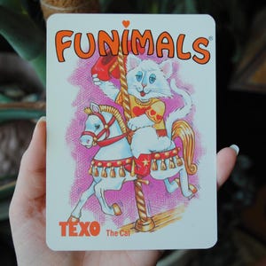 May include: A vintage FUNIMALS card featuring a white cat riding a carousel horse. The cat wears a yellow outfit with red accents and holds a red hat. The card has the text "FUNIMALS" and "TEXO The Cat".