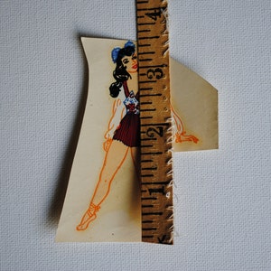 May include: A vintage paper doll cutout of a woman wearing a blue and white patterned dress and a blue bow in her hair. The doll is standing next to a ruler.
