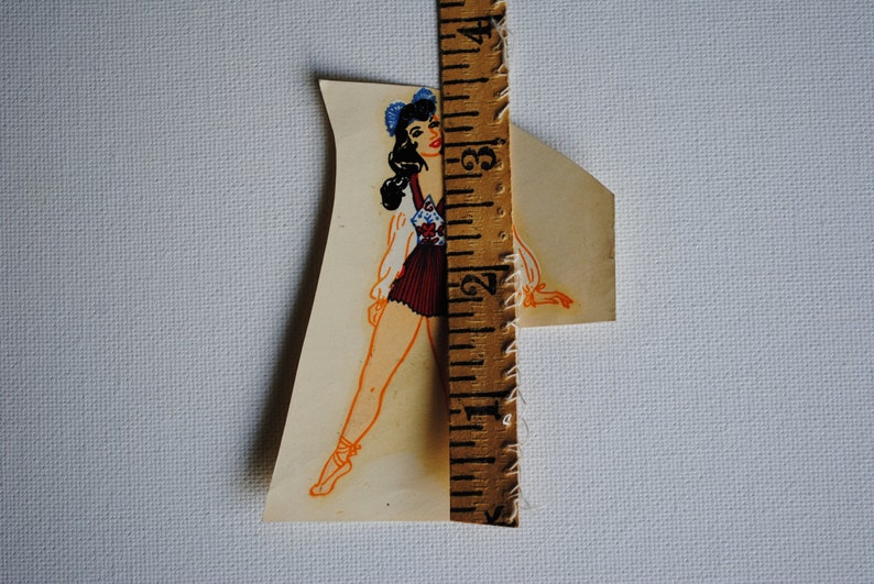 May include: A vintage paper doll cutout of a woman wearing a blue and white patterned dress and a blue bow in her hair. The doll is standing next to a ruler.