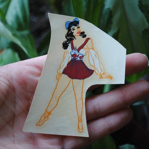 May include: A vintage paper doll cutout of a woman wearing a red and white dress with a blue and white floral pattern. She has dark hair and blue eyes and is wearing a blue bow in her hair.