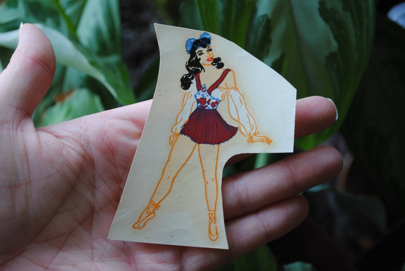 May include: A vintage paper doll cutout of a woman wearing a red and white dress with a blue and white floral pattern. She has dark hair and blue eyes and is wearing a blue bow in her hair.
