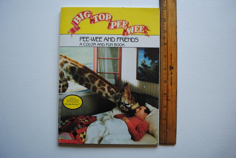 May include: A yellow and red book cover with the title "Big Top Pee-wee" and the subtitle "Pee-wee and Friends A Color and Fun Book". The cover features a giraffe reaching down to a man lying in bed.