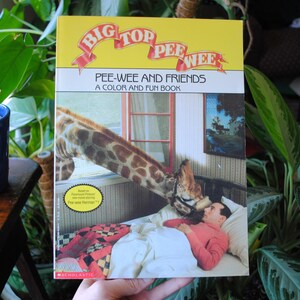 May include: A colorful book titled "Big Top Pee-wee: Pee-wee and Friends, A Color and Fun Book" features an illustration of a giraffe and a person in a bed. The book cover has a yellow background with red, white, and blue accents.
