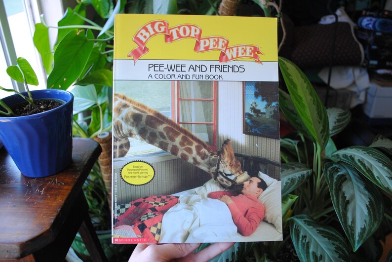 May include: A colorful book titled "Big Top Pee-wee: Pee-wee and Friends, A Color and Fun Book" features an illustration of a giraffe and a person in a bed. The book cover has a yellow background with red, white, and blue accents.