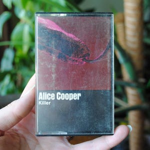 Puede incluir: Una cinta de casete con un diseño negro y rojo. El texto "Alice Cooper" y "Killer" está impreso en la parte delantera de la cinta.