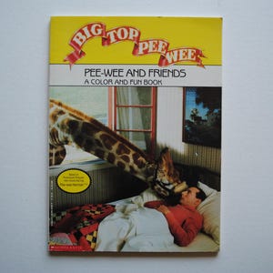 May include: A yellow and red book cover with the title "Big Top Pee-wee" and the text "Pee-wee and Friends A Color and Fun Book". The cover features a giraffe with a man lying on a bed in the background.