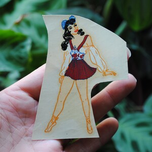 May include: A vintage paper doll cutout of a woman wearing a red and white dress with a blue floral pattern. She has dark hair and is wearing a blue bow on her head. The doll is posed with one leg extended and the other bent at the knee.