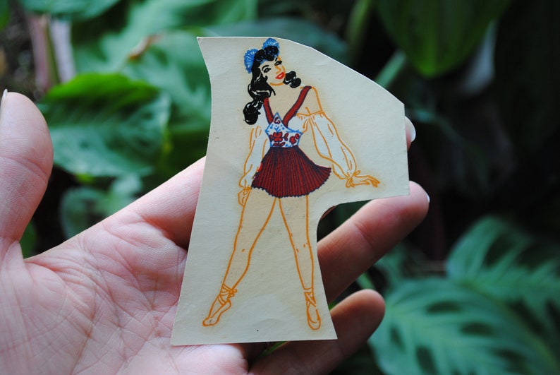 May include: A vintage paper doll cutout of a woman wearing a red and white dress with a blue floral pattern. She has dark hair and is wearing a blue bow on her head. The doll is posed with one leg extended and the other bent at the knee.