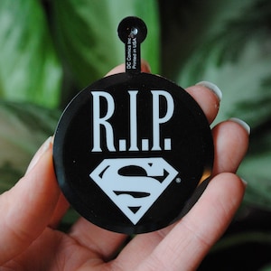 Puede incluir: Un botón negro con un logotipo de Superman blanco y el texto "R.I.P." en letras blancas.