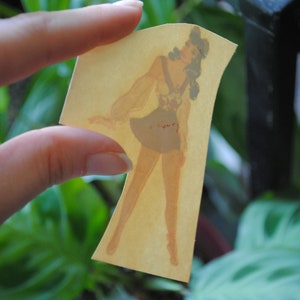 May include: A vintage paper cutout of a woman wearing a light brown dress with a red and white pattern. The cutout is on a yellowed paper background.