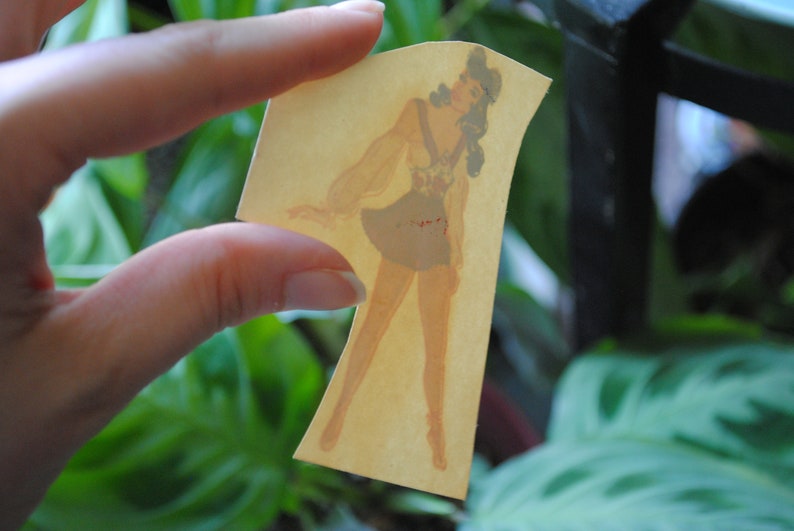 May include: A vintage paper cutout of a woman wearing a light brown dress with a red and white pattern. The cutout is on a yellowed paper background.
