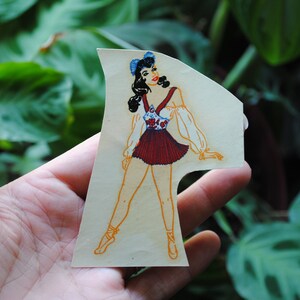 May include: A vintage paper doll cutout of a woman wearing a red and white dress with a blue flower pattern. She has dark hair and is wearing blue shoes.