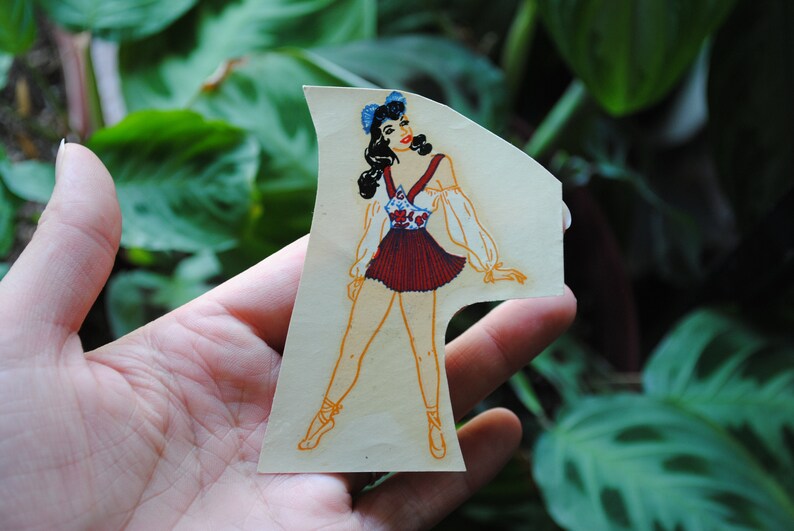 May include: A vintage paper doll cutout of a woman wearing a red and white dress with a blue flower pattern. She has dark hair and is wearing blue shoes.