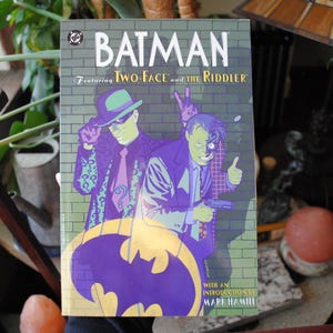 May include: A comic book cover featuring Batman, Two-Face, and the Riddler. The cover is illustrated in a colourful, graphic style. The title "Batman" is in large, bold letters. The subtitle reads "Featuring Two-Face and the Riddler". The cover also includes the text "With an introduction by Mark Hamill".