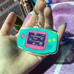 May include: A teal blue and pink Game Boy Advance keychain with glitter. The screen reads "Assman? Or Titsman?" and the bottom reads "Piss Boy Advance".