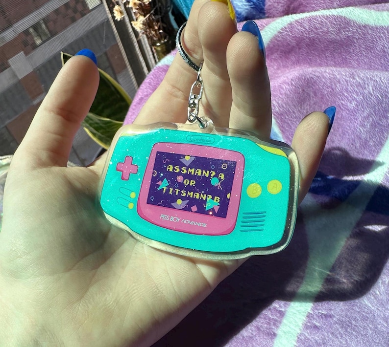 May include: A teal blue and pink Game Boy Advance keychain with glitter. The screen reads "Assman? Or Titsman?" and the bottom reads "Piss Boy Advance".