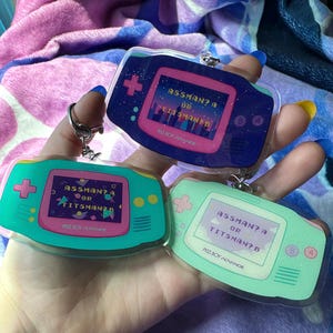 May include: Three clear plastic keychains shaped like a handheld game console. Each keychain has a pink and green screen with the text "ASSMAN? A OR TITSMAN? B" and the words "PISS BOY ADVANCE" at the bottom.