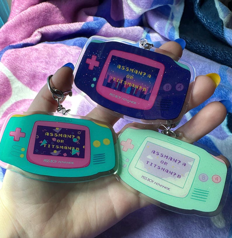 May include: Three clear plastic keychains shaped like a handheld game console. Each keychain has a pink and green screen with the text "ASSMAN? A OR TITSMAN? B" and the words "PISS BOY ADVANCE" at the bottom.