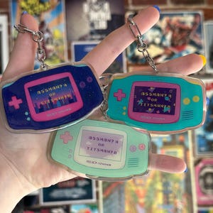 May include: Three blue, green, and light green Game Boy Advance keychain charms with a pink screen that reads "ASSMAN? A OR TITSMAN? B".