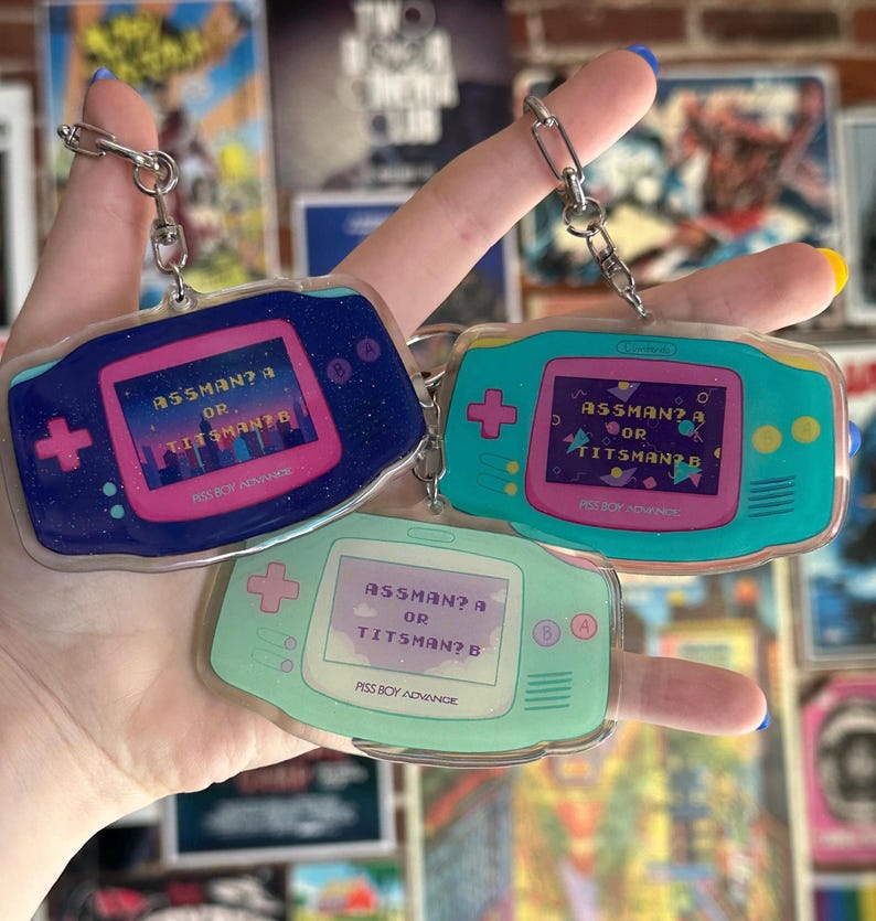 May include: Three blue, green, and light green Game Boy Advance keychain charms with a pink screen that reads "ASSMAN? A OR TITSMAN? B".