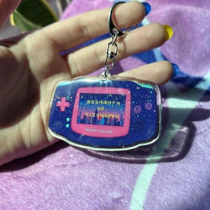 May include: Pink and blue Game Boy Color keychain with glitter accents. The screen displays the text "Assman? Or Git Shmantz?" and the words "Nintendo" and "Game Boy" are visible on the bottom of the keychain.