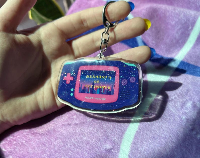 May include: Pink and blue Game Boy Color keychain with glitter accents. The screen displays the text "Assman? Or Git Shmantz?" and the words "Nintendo" and "Game Boy" are visible on the bottom of the keychain.