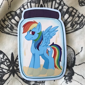 May include: A blue and rainbow pony with a lightning bolt symbol on its flank is trapped inside a glass jar. The pony has a sad expression and is surrounded by sand.