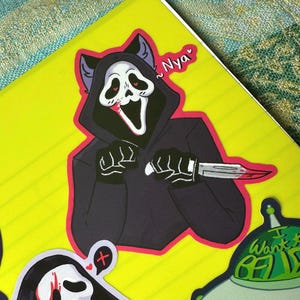 May include: A sticker of a black-hooded figure with a white mask and cat ears holding a bloody knife. The figure has red eyes and a wide, open mouth. The sticker has a pink border and the text "~Nyaa" is written in pink above the figure.